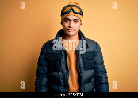 Junger brasilianischer Skifahrer Mann mit Schnee Sportbekleidung und Skibrille auf gelbem Hintergrund sieht schläfrig und müde, erschöpft von Müdigkeit und Kater, Stockfoto