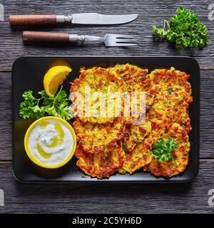 Tsatziki Sauce und Zwiebel Pfannkuchen serviert auf einem schwarzen Teller mit Zitrone auf einem dunklen Holzhintergrund mit Besteck, flach legen, close-up Stockfoto