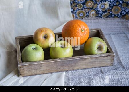 Stillleben mit Äpfeln und Orange in einer Holzkiste auf einem Hintergrund von Draperie. Stockfoto