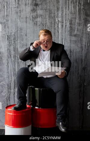 Schockiert fett kaukasischen Geschäftsmann überprüft Nachrichten über Ölreserven, er denkt, in Kontemplation, sitzt auf Fass Stockfoto