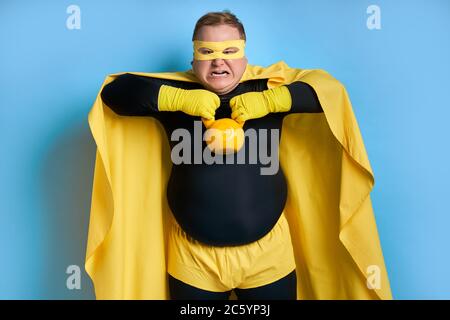 Starker fetter kaukasischer Superheld zeigt Muskeln an der Kamera, emotionaler Mann posiert mit Kettlebells isoliert auf blauem Hintergrund Stockfoto