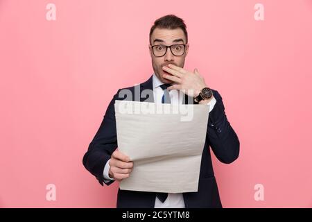 Überrascht elegantes Modell in Anzug Brille, Zeitung lesen und den Mund mit der Hand, auf rosa Hintergrund stehen Stockfoto