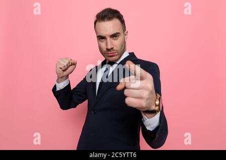 Verärgert junger Mann im Anzug hält Faust nach oben, zeigt Finger und bedrohlich, auf rosa Hintergrund stehen Stockfoto