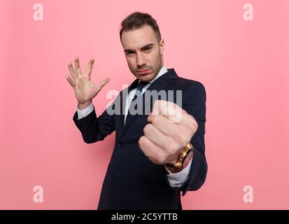 Junger eleganter Geschäftsmann im Anzug, der die Faust hochhält und stanzt, auf rosa Hintergrund schlägt und steht Stockfoto