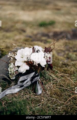 brautstrauß aus weißen Rosen, Lilie, Burgunder Nelken, protea, getrockneten Zweigen, Brunia und Leukadendron mit grauen und schwarzen Bändern in der Nähe des getrockneten Grases Stockfoto