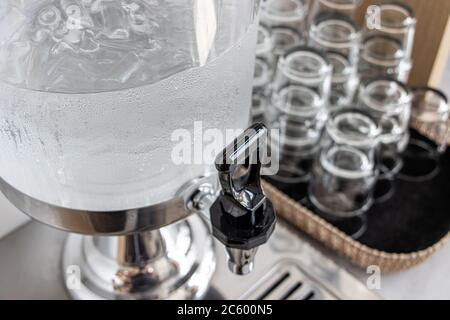 Trinkwasser mit Eis in einem Selbstbedienungswasserspender mit Glas auf dem Tisch reinigen. Wasserhahn aus Glasbehälter für kaltes Wasser am Buffet Self Service Stockfoto