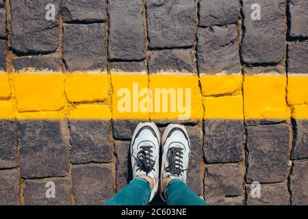 Damenfüße in weißen Sneakers stehen auf dem Bürgersteig hinter der gelben Linie, Körperteile überqueren die Linie nicht. Stockfoto