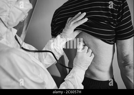 Ausschnitt aus der Nahaufnahme eines Arztes in einem Schutzanzug, der ältere männliche Patienten zu Hause mit Phonendoskop untersucht und seinen Rücken überprüft, schwarz-weiß. Coronavirus-Konzept Stockfoto