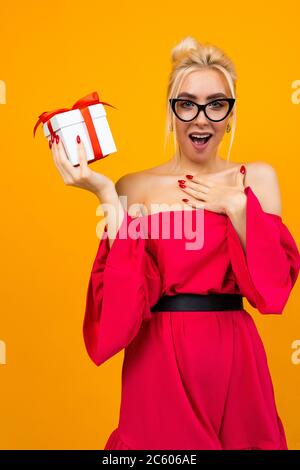 Angenehm überrascht attraktive junge Frau in einem roten Kleid erhielt eine weiße Geschenkbox mit einem roten Geburtstagsband auf einem orangen Hintergrund Stockfoto