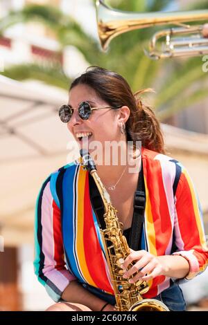 Glücklich aussehende junge schöne Frau trägt eine Brille und hält ein goldenes Saxophon auf einem unscharf Hintergrund. Street Music und Lifestyle Konzept. Stockfoto