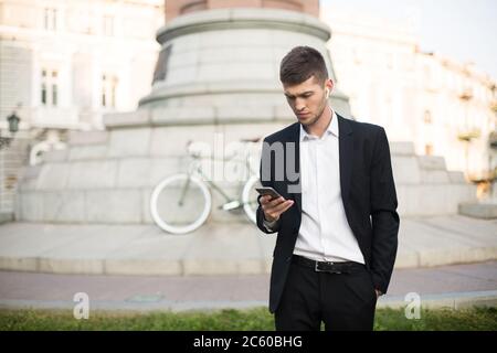 Junge ernsthafte Geschäftsmann in klassischen schwarzen Anzug und weißen Hemd mit drahtlosen Ohrhörern nachdenklich mit Handy mit Retro-Fahrrad auf Fuzzy Stockfoto