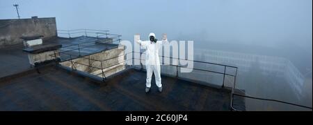 Panorama-Schnappschuss eines Mannes in weißer Overall und Gasmaske, der mit den Händen nach oben auf dem Dach eines Hochhauses steht und das Stoppschild in Quarantäne sticht Stockfoto