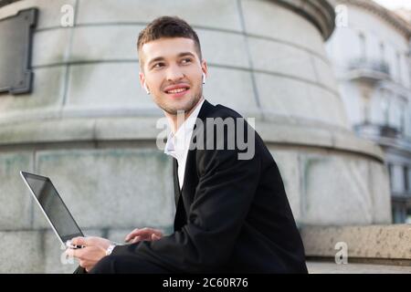 Junger lächelnder Mann in klassischem schwarzen Anzug mit kabellosen Ohrhörern schaut glücklich mit Laptop in den Händen zur Seite, während er Zeit im Freien verbringt Stockfoto