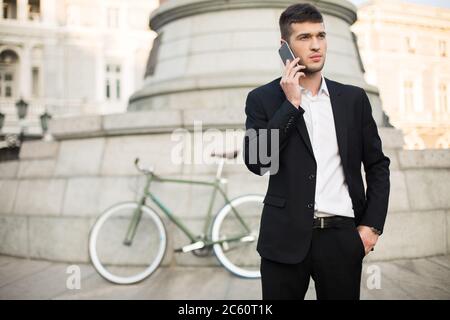 Junge, gutaussehende Geschäftsmann in klassischem schwarzen Anzug und weißem Hemd reden auf Handy, während nachdenklich neben mit Retro-Fahrrad auf Stockfoto