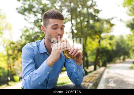 Junge attraktive Mann in blauem Hemd sitzen auf Bank glücklich essen Sandwich während die Zeit in schönen Park Stockfoto