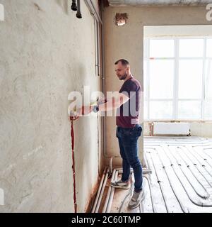 Junger Mann in leerer unvollendeter Wohnung, die die Wand mit Hilfe von Maßband misst, steht auf Fußbodenheizung Rohrsystem gegen Panoramafenster. Konzept der Renovierung und Renovierung. Stockfoto