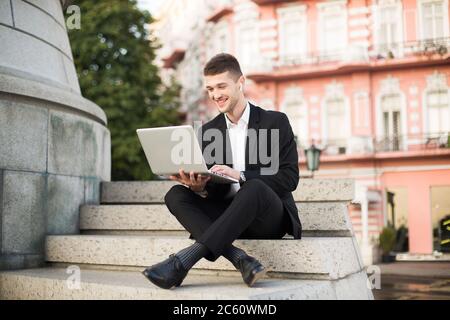 Junger fröhlicher Geschäftsmann in klassischem schwarzen Anzug mit kabellosen Kopfhörern, der gerne mit einem Laptop auf kleinen Treppen sitzt und Zeit verbringt Stockfoto