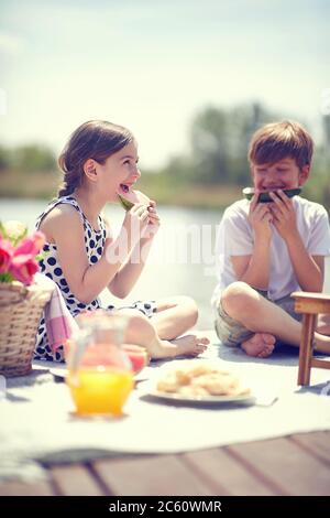 Kinder auf dem Holzsteg essen Wassermelone. Sommerfreude, nettes Mädchen essen frische Wassermelone auf dem Pier. Stockfoto