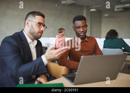 Portrait eines reifen bärtigen Geschäftsmannes, der mit einem jungen afrikanischen Kollegen spricht, während er im Büro über ein Projekt und einen Kopierraum spricht Stockfoto