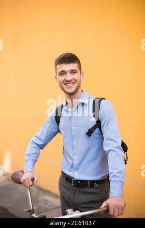 Junger lächelnder Mann in blauem Hemd und Rucksack, der mit dem Fahrrad fröhlich neben dem orangen Hintergrund im Freien steht Stockfoto