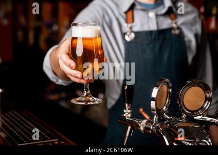 Bier am freitagabend. Der Barkeeper gibt ein Glas helles Bier mit Schaum im dunklen Innenraum Stockfoto