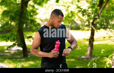 Afroamerikanischer Läufer mit einer Flasche Wasser, der nach dem Training im Park seine Smartwatch überprüft Stockfoto