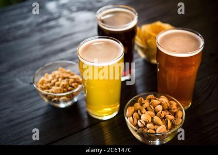 Gläser mit drei verschiedenen Biersorten. Nüsse, Zwieback und Pommes in Schüsseln auf Holztisch Stockfoto