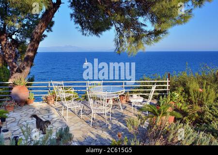 Picknicktisch und Stühle unter Pinien im Innenhof an der Küste der Ägäis bei Sonnenuntergang, weiße Segelyacht im Hintergrund. Warmes Abendlicht. A Stockfoto