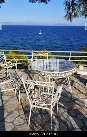 Picknicktisch und Stühle unter Pinien im Innenhof an der Küste der Ägäis bei Sonnenuntergang, weiße Segelyacht im Hintergrund. Warmes Abendlicht. A Stockfoto