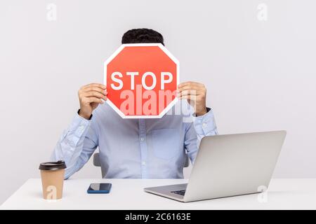 Mann Mitarbeiter sitzt im Büro Arbeitsplatz mit Laptop, versteckt Gesicht hinter Stopp-Symbol, Warnung mit roten Verkehrszeichen, Symbol der Verbotsbeschränkung