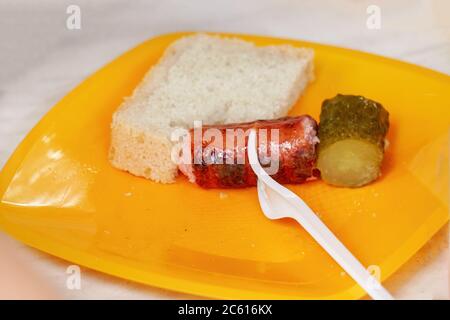 Gegrillte Wurst auf einer Gabel, Gurke und Brot auf einem gelben Kunststoffteller. Einweggeschirr Stockfoto