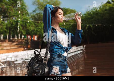 Portrait of beautiful lady in denim shirt and shorts standing with black backpack and drinking water under rain in park Stockfoto