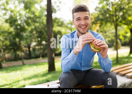 Junger lachender Mann in blauem Hemd sitzt auf der Bank mit Sandwich in den Händen freudig in der Kamera mit gemütlichen Park auf dem Hintergrund zu suchen Stockfoto