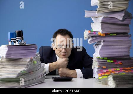 Müder Büroangestellter legt seinen Kopf auf die Fäuste, inmitten von hohen Dokumentenstapeln Stockfoto