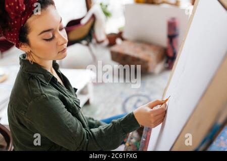 Junge ernsthafte Frau mit dunklen lockigen Haaren sitzt auf Stuhl nachdenklich Bild auf Leinwand in der modernen Kunst-Werkstatt allein zeichnen Stockfoto