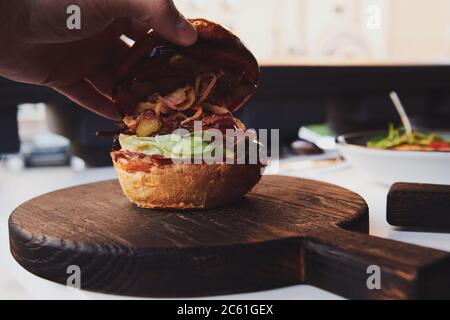 Die Hand des Mannes öffnet den Burger. Burger mit Entenfleisch auf dem Holzbrett im Restaurant. Frisches Brötchen mit Salat und eingelegter Gurke. Stockfoto