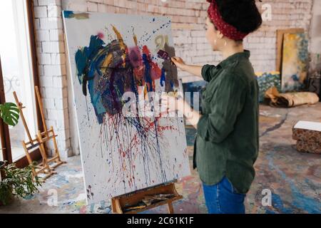 Junge hübsche Frau mit dunklen lockigen Haaren traumhaft Zeichnung Bild auf Leinwand mit hellen Farben während der Zeit in großen gemütlichen Werkstatt Stockfoto