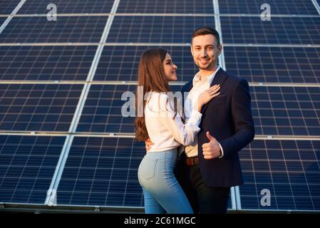 Junges und schönes Paar steht dicht beieinander durch ein Solarpanel, Mann zeigt Daumen hoch, Seitenansicht Stockfoto