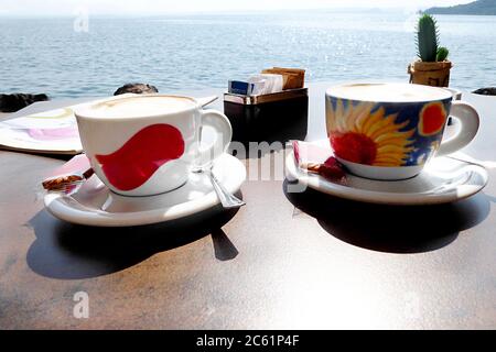 Zwei Tassen und Untertassen mit Kaffee Cappuccino Getränk mit kleinen Keksen und Teelöffel, draußen auf einem Tisch mit Blick auf den Gardasee glitzernden in den Sonnen Stockfoto