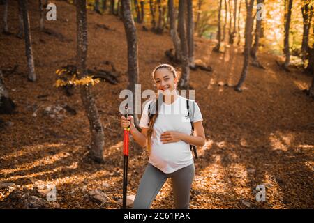 Portrait einer jungen, glücklichen schwangeren Frau nordic Walking im Herbstwald mit Rucksack und Trekkingstöcken. Gesund, Wellness und aktive Lebensweise in der Mutterschaft. Stockfoto