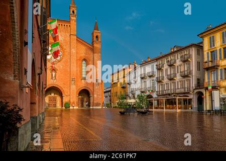 Nasser gepflasterter Stadtplatz, alte Häuser und Kathedrale San Lorenzo, die von Sonnenlicht unter regnerischem Himmel in Alba, Piemont, Norditalien beleuchtet wird. Stockfoto