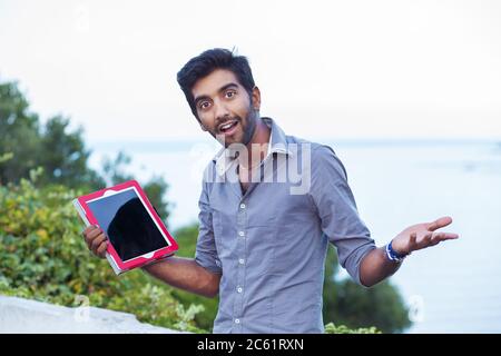 Ein lächelnder Mann, Student zeigt Gleichgültigkeit, also was Geste mit Händen halten zeigt einen Tablet-Pad-Bildschirm, während vor einem Stadtpark Witz stehen Stockfoto