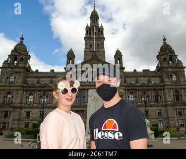 Glasgow, Schottland, Großbritannien. Juli 2020. Im Bild: Ein Paar sah, wie sie ihre Sommerkattire auf dem George Square schaukelte, mit einer Dame in einer Sonnenbrille mit Gänseblümchen-Muster und einem Herrn mit schwarzer Gesichtsmaske und schwarzer Baseballmütze, mit den City Chambers als Hintergrund. Menschen auf dem George Square. Bars und Restaurants werden in Glasgow wieder eröffnet, da die Sperrungen leichter werden. Von heute an begrüßen Pubs, Bars, Cafés und Restaurants in England, Schottland und Nordirland zum ersten Mal seit Beginn der Sperre im März Kunden. Quelle: Colin Fisher/Alamy Live News Stockfoto