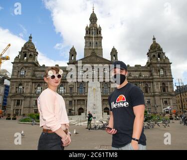 Glasgow, Schottland, Großbritannien. Juli 2020. Im Bild: Ein Paar sah, wie sie ihre Sommerkattire auf dem George Square schaukelte, mit einer Dame in einer Sonnenbrille mit Gänseblümchen-Muster und einem Herrn mit schwarzer Gesichtsmaske und schwarzer Baseballmütze, mit den City Chambers als Hintergrund. Menschen auf dem George Square. Bars und Restaurants werden in Glasgow wieder eröffnet, da die Sperrungen leichter werden. Von heute an begrüßen Pubs, Bars, Cafés und Restaurants in England, Schottland und Nordirland zum ersten Mal seit Beginn der Sperre im März Kunden. Quelle: Colin Fisher/Alamy Live News Stockfoto