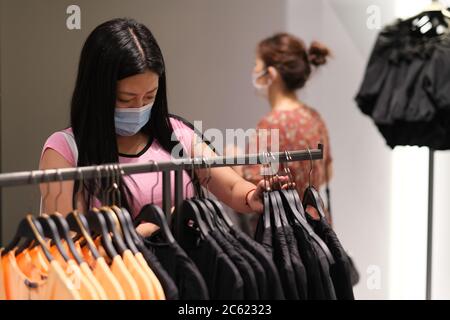 Asiatische junge Frau in Gesichtsmaske, um Coronavirus zu verhindern, Auswahl Tuch in Bekleidungsgeschäft. Hintergrund verwischen Stockfoto