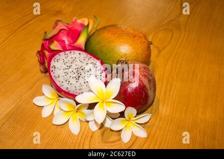 Eine Sammlung exotischer und tropischer Früchte, nämlich Drachenfrucht, Mango und Granatapfel, arrangiert mit Frangipani Blumen, auf einem hölzernen Hintergrund Stockfoto