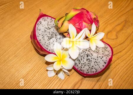 Eine geschnittene und ganze Drachenfrucht, auch als Pitaya bekannt, arrangiert mit Frangipani-Blumen auf einem hölzernen Hintergrund Stockfoto