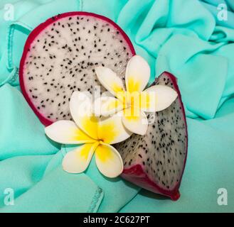 Eine geschnittene Drachenfrucht, auch als Pitaya bekannt, arrangiert mit Frangipani-Blumen auf einem türkisfarbenen Tuch Stockfoto