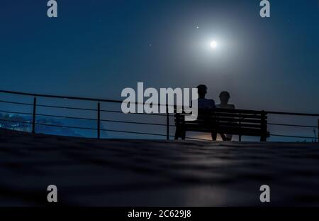 Silhouette eines liebevollen Paares auf einer Bank im Park auf dem Hintergrund des Mondes und Mondpfad auf Wasseroberfläche. Romantischer Abend auf dem Meer unter Stockfoto