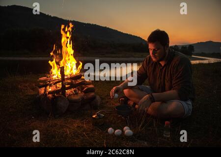 Mann sitzt neben Lagerfeuer und kocht Spiegeleier Stockfoto